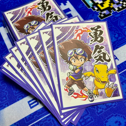 A fan of illustrated trading cards or sticker cards showing a chibi anime character with goggles and a yellow dinosaur-like companion, photographed on a blue patterned surface.