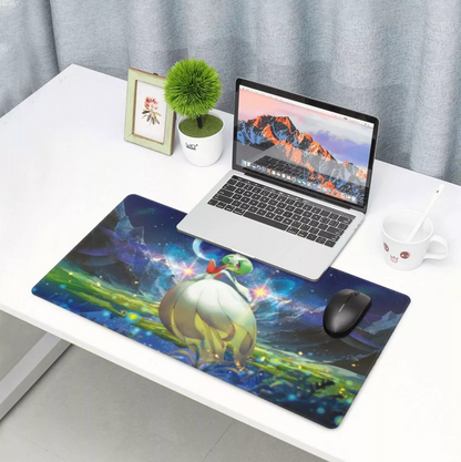 A large rectangular gaming desk mat with a colorful fantasy creature illustration lies on a white desk, alongside an open laptop, mouse, pencil, small plant, and framed picture. The scene is set in a bright room with gray curtains in the background.