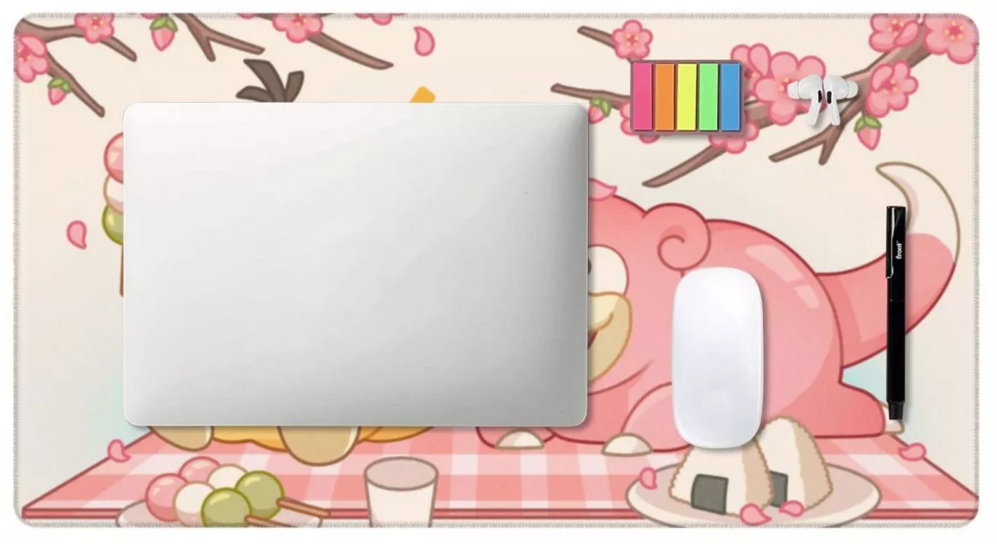 A rectangular desk mat with a pink and beige cherry blossom picnic illustration, shown with a silver laptop, white mouse, and black stylus on top against a white background.