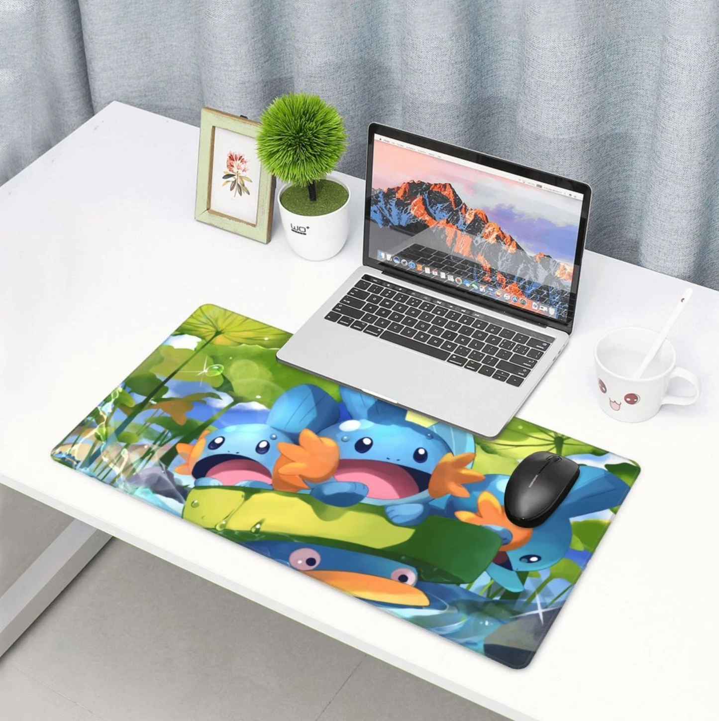 A large rectangular desk mat with a colorful cartoon fish illustration sits on a white desk next to an open laptop, a small potted plant, a framed picture, and a white mug.