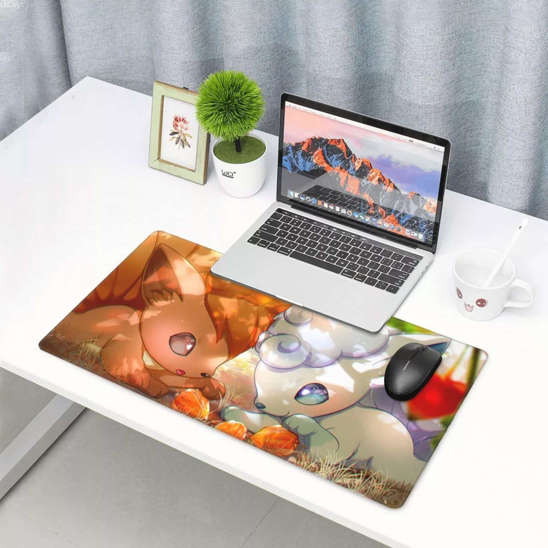 A large rectangular desk mat with colorful anime-style character artwork is placed on a white desk, alongside an open laptop, mouse, plant, mug, and small framed picture. The desk mat is the product being shown in a real home-office setting, so this is Lifestyle rather than Product/Packaging.