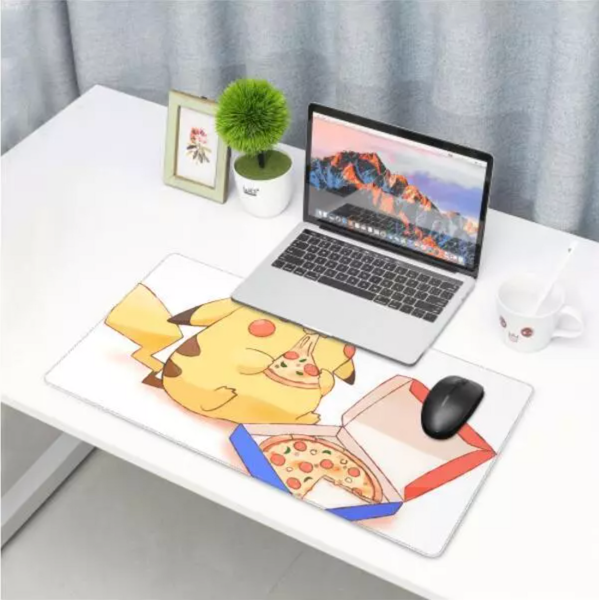 A large desk mat featuring a cartoon Pikachu holding a slice of pizza next to an open pizza box, shown on a white desk with a laptop, mouse, mug, and small plant in a home office setting.