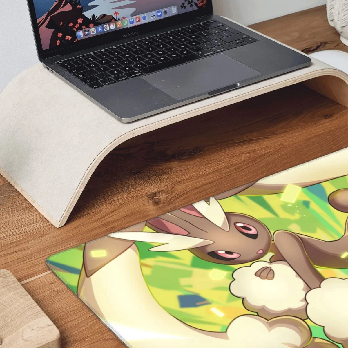 A wooden desk organizer shelf holding an open laptop, with a colorful printed desk mat featuring a cartoon rabbit character on a wood-grain desk surface.