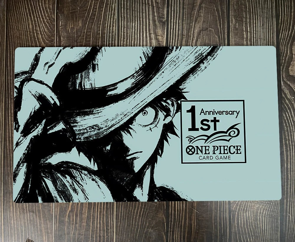 A rectangular One Piece Card Game playmat with a black and white illustration of Luffy on a light blue background, placed on a dark wood surface. The playmat features a '1st Anniversary ONE PIECE CARD GAME' logo on the right side.