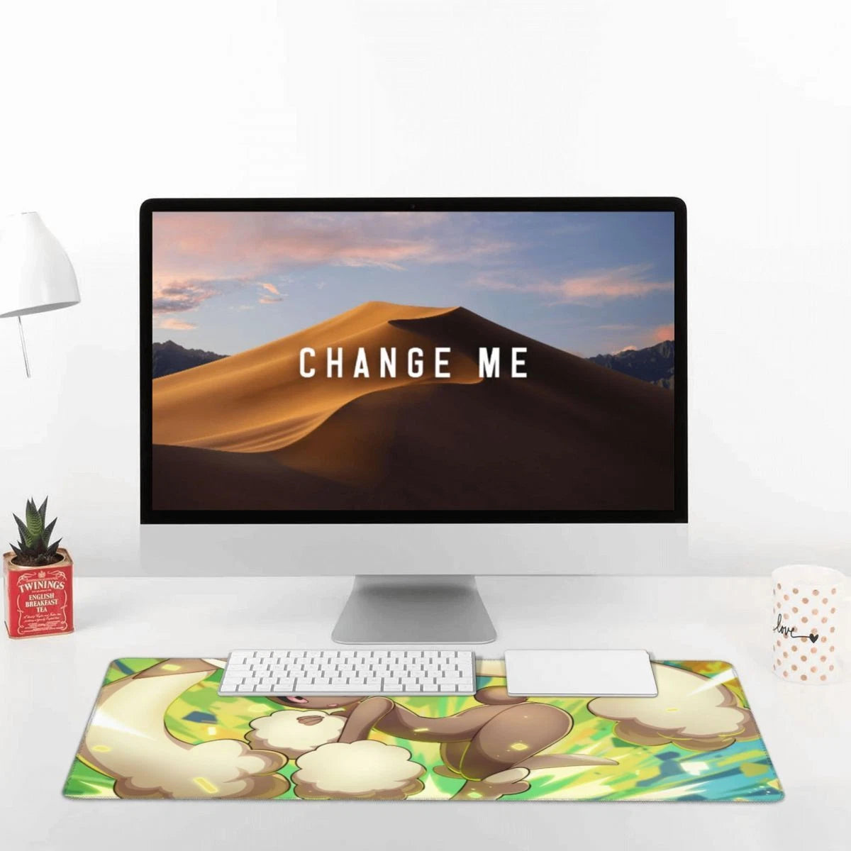 A large desk mat with a colorful cartoon-style illustration sits on a white desk in front of an iMac. The mat features a whimsical scene with cream-colored creatures on a green and yellow background.