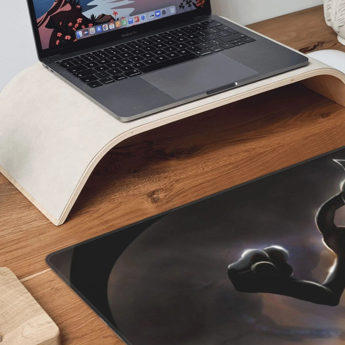 A curved wooden desk riser holding an open laptop, placed on a wood-grain desk with a large dark printed mat in the foreground.