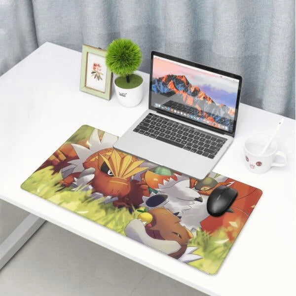 A large desk mat featuring colorful Pokémon characters, shown on a white desk alongside a laptop, mouse, plant, and mug.